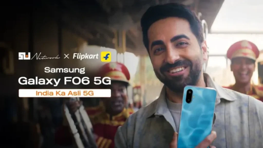 The image highlights the tagline 'India Ka Asli 5G,' with the actor holding the phone in a vibrant blue finish, smiling, while a festive band plays in the background. The campaign is part of Samsung's viral Eyebrow Dance Challenge, engaging users in a fun and interactive way to promote the Galaxy F06 5G